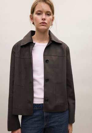 Jeune femme portant une veste grise foncée boutonnée sur un haut blanc et un jean bleu sur un fond clair uni.