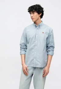 Ανοιχτό μπλε ριγέ πουκάμισο με κουμπιά και μακριά μανίκια, με κεντητό λογότυπο ζώου στο στήθος. Φορεμένο με ανοιχτόχρωμο τζιν παντελόνι.