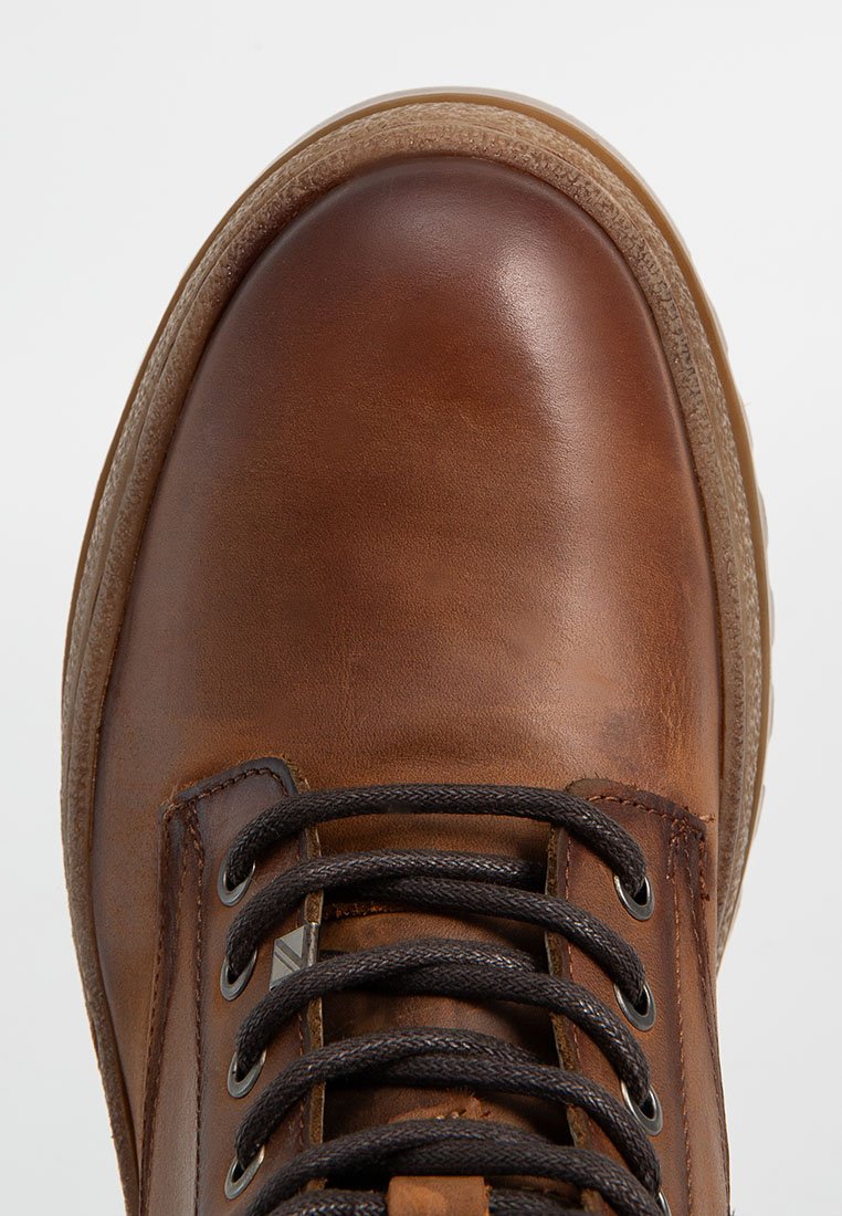 Bota de cuero marrón con punta redondeada, superficie texturizada, cordones negros y suela de goma. Los acentos incluyen ojales de metal y costuras.