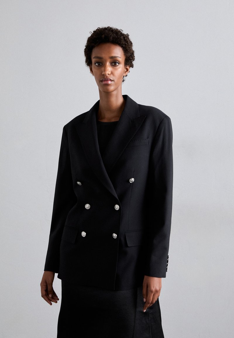 Zwarte dubbelzijdige blazer gemaakt van gladde stof met zilveren knopen, notch revers en zakken aan de voorkant. Model staat tegen een lichte achtergrond.
