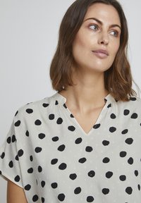 Blusa leggera in crema con motivo a pois neri sparsi, scollatura a V, maniche corte e dettagli in tessuto testurizzato discreti.