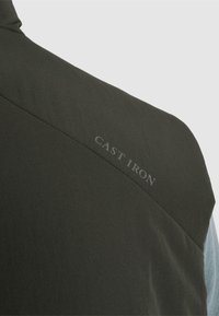 Épaule de veste vert foncé avec couture et texte de marque "CAST IRON" estompé sur la partie haute du dos.