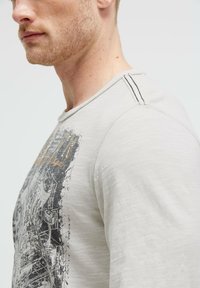 Helles graues Langarmshirt mit einem grafischen Druck in Schwarz und Gold, mit akzentuierten Nähten an der Schulter und einer lockeren Passform.