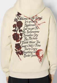 Sudadera crema con gráficos de flores rojas y texto en la parte trasera, con un detalle de calavera. Puños y dobladillo acanalados, textura de tela suave.