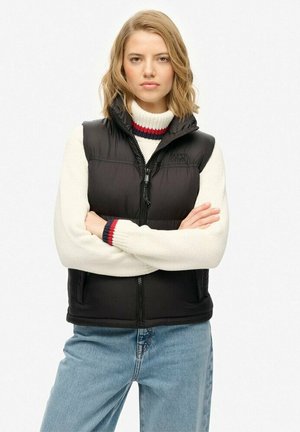 Young woman with blonde hair wearing a black puffer vest over a white sweater with red and blue stripes, standing with arms crossed.