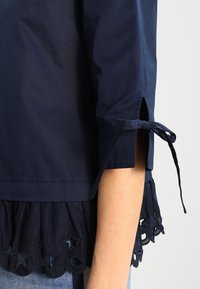 Marinblus i bomull med en utsvängd nederkant och stjärn- och cirkelutskärningar samt knytningar på ärmarna, som visar en mjuk textur och skräddarsydd passform.