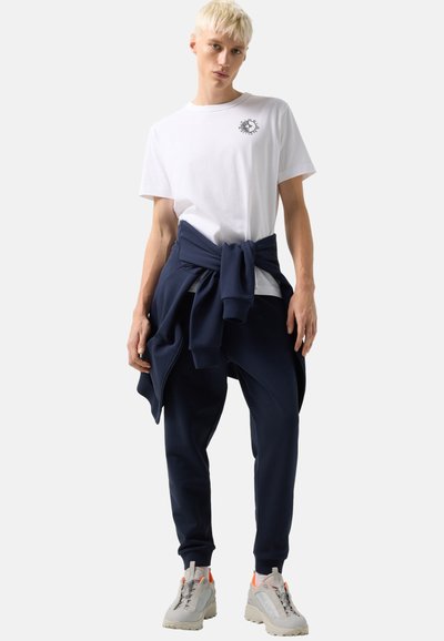 Mladá osoba s krátkými blond vlasy, oblečená v bílém tričku s potiskem, tmavě modré mikině uvázané kolem pasu, tmavě modrých teplácích a béžových teniskách.