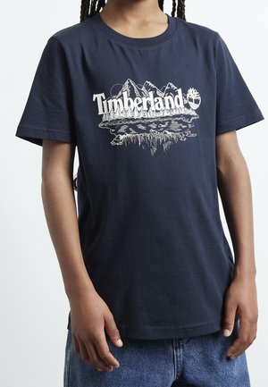 Person iført en navyblå Timberland t-shirt med en hvid grafik af bjerg og skov og jeans, hænderne afslappet ned langs siderne.