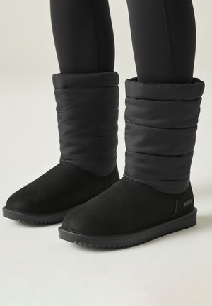 Schwarze gepolsterte Winterstiefel, getragen mit schwarzen Leggings auf hellem Hintergrund, zeigen Vorder- und Seitenansichten beider Füße.