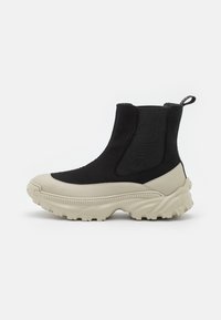 Botas de tobillo negras y crema con parte superior de tejido elástico, suela de goma y diseño de suela gruesa, que cuentan con una pestaña de tirón en la parte posterior.