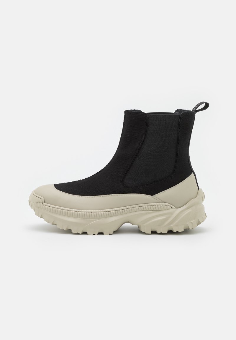 Botas de tobillo negras y crema con parte superior de tejido elástico, suela de goma y diseño de suela gruesa, que cuentan con una pestaña de tirón en la parte posterior.