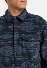 Camicia in pile con tonalità scure e motivi a rombi. Presenta una chiusura con bottoni, colletto e taschino sul petto. Tessuto strutturato.