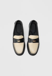 Schwarz-cremefarbene Leder-Loafer mit abgerundetem Zehenbereich, ausgestattet mit einem dekorativen Riemen und Metallakzent. Glatte Textur und kontrastierende Farben.