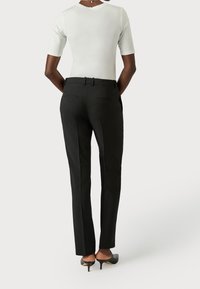 Person wearing black tailored pants, white short-sleeve top, and black heeled shoes, standing facing away with hands behind back.