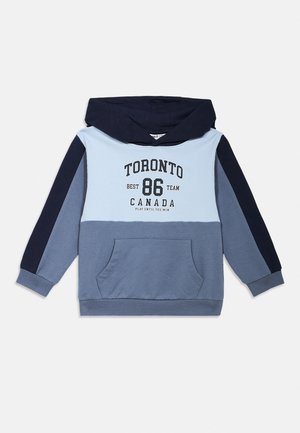 Hūdija ar gaiļa kapuci gaiši zilu, pelēku un tumši zilu paneļu kombinācijā, tumši zils kapucis, priekšējā kabata un teksts „Toronto 86 Canada“ uz krūtīm.