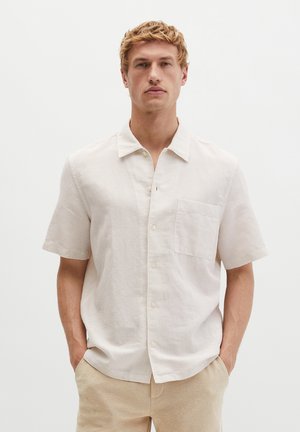 Blonder Mann trägt ein kurzärmeliges, hellbeiges Hemd mit Brusttasche und beigen Hosen, steht mit den Händen in den Taschen.