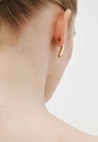 Pendientes en tono dorado con un diseño liso y alargado, y un pequeño pendiente con un acento floral, llevados cerca de la oreja en vista de perfil.