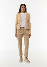 Beige gerippter Cardigan über einem hellgrauen T-Shirt, kombiniert mit beigen Cargo-Hosen und beigen Sneakers mit dicken Sohlen. Einfache Knopfdetails sind sichtbar.
