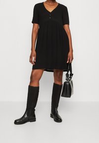 Vestido negro de manga corta con escote en V y detalles de botones. El material es ligero y fluido, combinado con botas de cuero negras hasta la rodilla y un bolso.