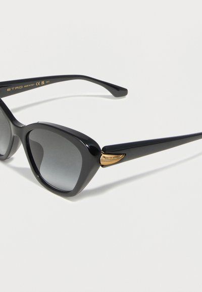 Lunettes de soleil noires style œil de chat avec des verres dégradés foncés et des détails dorés près des tempes, présentées sur un fond clair.