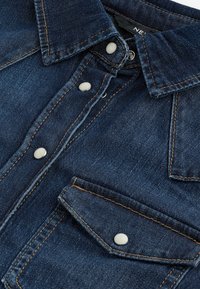 Camicia di jeans blu scuro con colletto classico, bottoni a scatto e tasca sul petto. Presenta cuciture a contrasto color cuoio. Texture liscia.