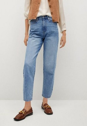 Vrouw draagt lichtblauwe jeans met hoge taille en rechte pijpen, beige blouse, bruine mouwloze buttoned vest en bruine loafers met gouden kettingdetail.