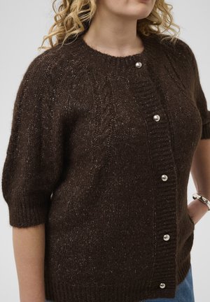 Femme portant un cardigan tricoté marron foncé avec des boutons en perle et des manches aux coudes, debout avec une main sur la hanche.
