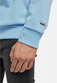 La mano destra appoggiata al fianco, indossando un felpa azzurro chiaro con un piccolo logo a righe rosso, bianco e blu sul polsino, abbinata a pantaloni neri.