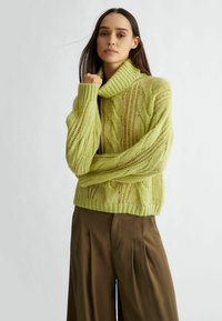 Maglione lavorato a maglia di colore verde lime, oversize, con un colletto alto e motivo a trecce; polsini e orlo a coste; indossato con pantaloni larghi marroni.