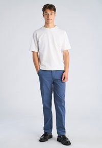 T-shirt bianca in cotone, vestibilità comoda, abbinata a pantaloni blu in cotone, taglio dritto, indossata con scarpe nere in pelle, in piedi su uno sfondo semplice.