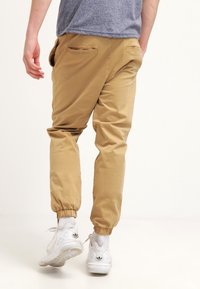 Osoba w beżowych spodniankach z podwijanym dołem, białych trampkach i szarym t-shircie, przedstawiona idąca w stronę wyjścia na białym tle.