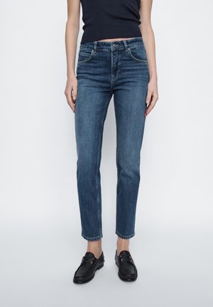 Vrouw draagt mid-rise slim fit blauwe jeans en zwarte loafers, staand tegen een effen lichte achtergrond.