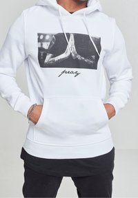 Witte hoodie met een afbeelding op de voorkant van handen in een biddende houding, met zichtbare tatoeages. Zwarte letters eronder luiden "pray".