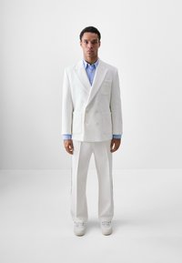 Weißer, doppelt gestalteter Anzugjacke mit großen vorderen Taschen, kombiniert mit weißen Hosen mit seitlichen Streifen und einem hellblauen Hemd darunter.