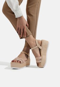 Sandali piattaforma beige con una cinghia intrecciata testurizzata, fibbia alla caviglia e suola in gomma bianca; abbinati a pantaloni marroni sartoriali.