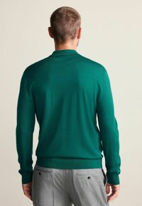 Grüner Strickpullover mit langen Ärmeln, rundem Ausschnitt und gerippten Bündchen, getragen zu grauer Hose. Glatte Textur, lässiges Design.