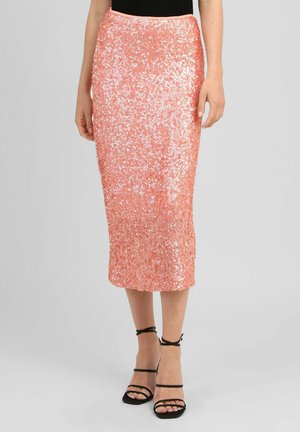 Femme portant une jupe midi à sequins couleur pêche associée à des sandales noires à talons hauts et brides, debout devant un fond uni.