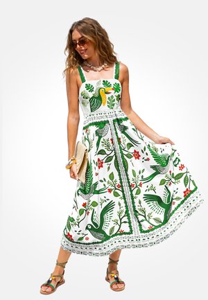 Frau trägt ein weißes Sommerkleid mit grünen Vogel- und Blattmustern, hält den Rock und eine beige Clutch, accessoriert mit Muschelhalskette und Sonnenbrille.