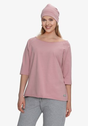 Uśmiechnięta kobieta, ubrana w różową, luźną bluzkę z dekoltem typu łódka i rękawami do łokcia, dopasowaną różową czapkę i jasnoszare spodnie dresowe, stojąca.