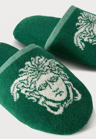 Πράσινες παντόφλες από πετσετέ ύφασμα με ένα περίτεχνο λευκό σχέδιο κεφαλής Μέδουσας στην επάνω επιφάνεια.