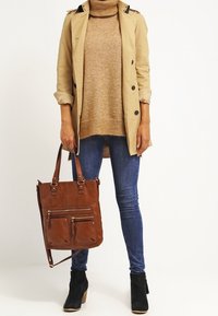 Manteau beige, pull à col roulé beige, jeans bleu foncé, bottines noires montantes et un sac fourre-tout en cuir marron avec plusieurs poches zippées.