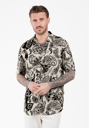 Homme aux cheveux courts et bruns avec des tatouages, portant une chemise à manches courtes à motif cachemire noir et blanc et un pantalon blanc, debout les mains jointes.