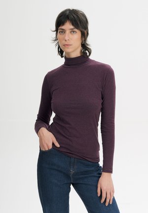 Jonge vrouw met kort donker haar die een paarse coltrui en blauwe spijkerbroek draagt, staand met één hand in de zak tegen een witte achtergrond.