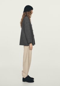 Blazer in lana grigia con finitura texturizzata, bottoni laterali, abbinato a pantaloni in lana color beige e stivaletti neri, su uno sfondo neutro.