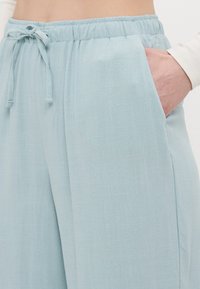 Světle modré kalhoty s širokými nohavicemi vyrobené z měkké, strukturované látky. Mají elastický pas se šňůrkou a šikmé boční kapsy.