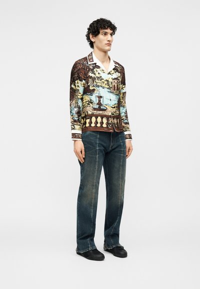 Νέος άνδρας που φοράει μακρυμάνικο πουκάμισο με εκτυπωμένες λεπτομέρειες τοπίου, ξεβαμμένα μπλε τζιν και μαύρα παπούτσια, στέκεται μπροστά σε απλό φόντο.