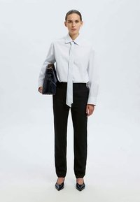 Woman standing in black pants, white shirt with matching long tie, holding a black handbag, wearing black pointed shoes, neutral background.