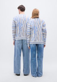 Πλεκτό πουλόβερ με μπλε και μπεζ μοτίβο με υφή κυματοειδών γραμμών· συνδυασμένο με ανοιχτό μπλε τζιν παντελόνι με φαρδιά μπατζάκια. Πίσω όψη δύο μορφών.