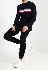 Marinblå tröja med ett horisontellt randmönster i blått, rött och grått; kombinerad med svarta joggingbyxor och vita sneakers, sittande på en pall.