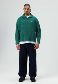 Turquoise fleece trui met een kraag, voorzien van lange mouwen en een casual pasvorm. Draagt over een wit T-shirt en gecombineerd met donkere spijkerbroek.